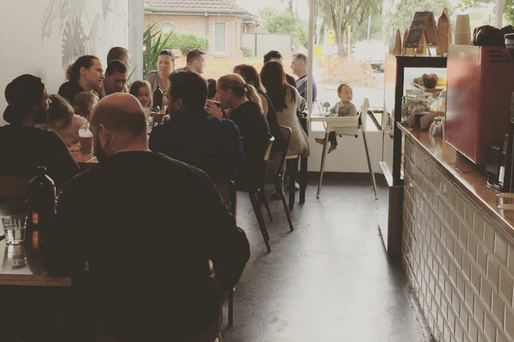 People sitting at tables inside a cafe with large windows showing an outdoor view.